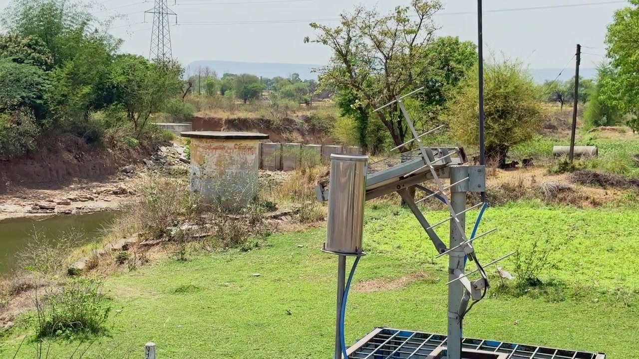 Primo piano di apparecchiature di monitoraggio alimentate a energia solare e installazione di antenna vicino alla sponda di un fiume rurale, circondato da vegetazione verde e paesaggio arido!