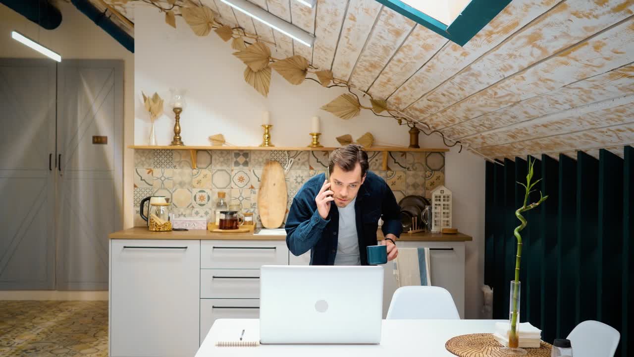 un joven trabajador independiente está hablando por teléfono en la cocina. resolve problemas de trabajo. trabaja en casa.