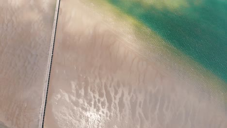 Slow-upwards-tilt-to-reveal-a-long-wooden-pier-stretching-across-sand-banks-of-land-into-the-vast-tropically-blue-ocean-featuring-islands-of-sand-below-the-gorgeous-blue-sky-featuring-fluffy-clouds