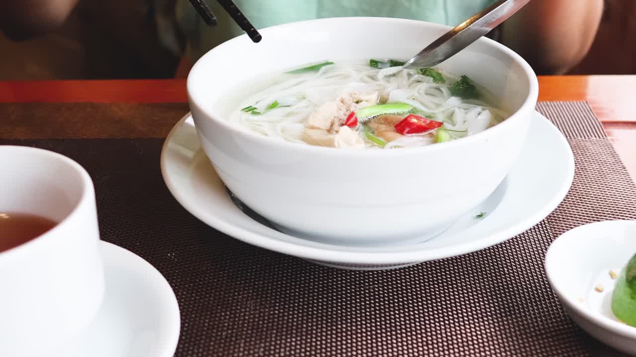 Close-up of a person using chopsticks and spoon to eat noodles with a cup of tea nearby.