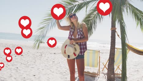 Multiple-heart-icons-floating-against-caucasian-woman-with-hat-adjusting-her-hair-on-the-beach