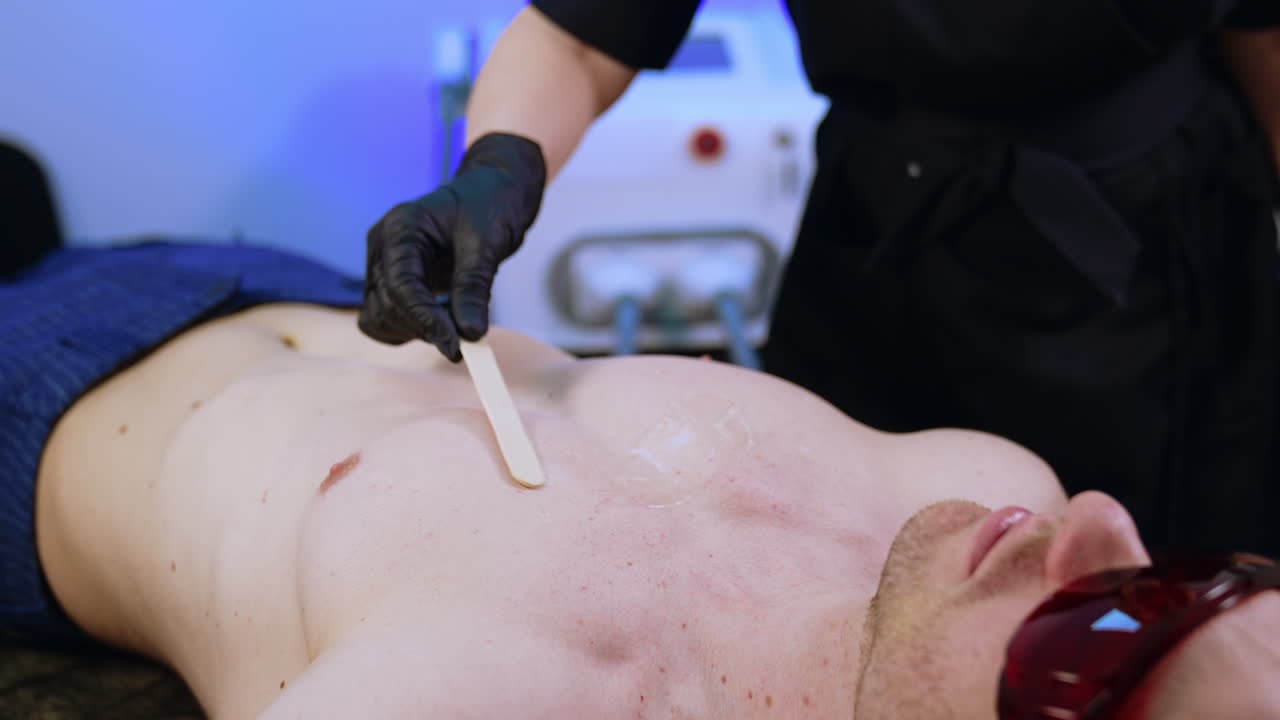 Caucasian male wearing protective goggles lies on the salon couch. Hands in black gloves apply transparent gel on man's chest.