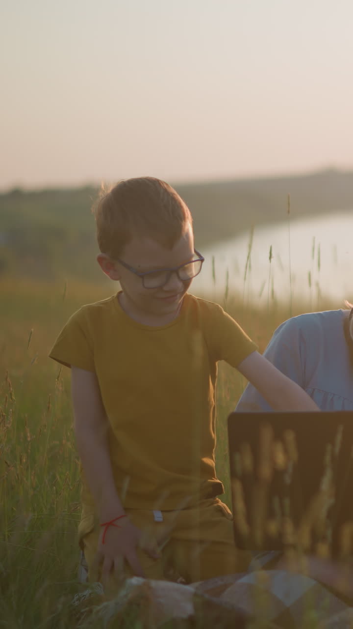 una mujer con una túnica azul se sienta en una bufanda en un campo cubierto de hierba, trabajando en una computadora portátil durante la puesta de sol. a su lado, un niño joven vestido de amarillo se sienta, mientras que otro niño de blanco se inclina a su lado