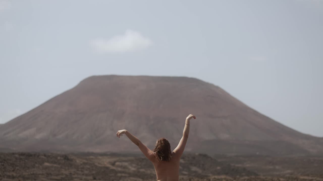 naked woman dancing and doing morning yoga in front of a massive volcano in the middle of the desert in Fuerteventura, canary islands
