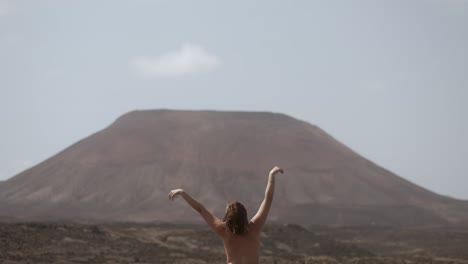 naked woman dancing and doing morning yoga in front of a massive volcano in the middle of the desert in fuerteventura, canary islands