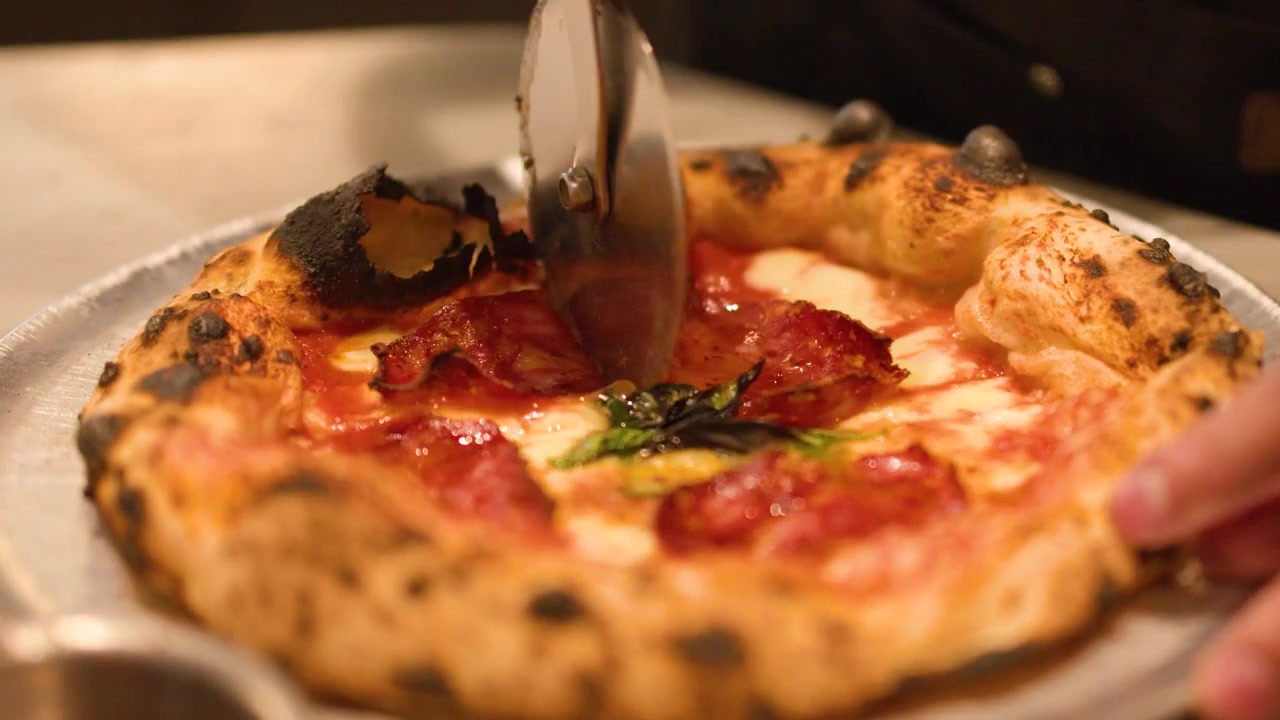 Primo piano di mani che tagliano una pizza calda al salame piccante su un vassoio di metallo usando una rotella.