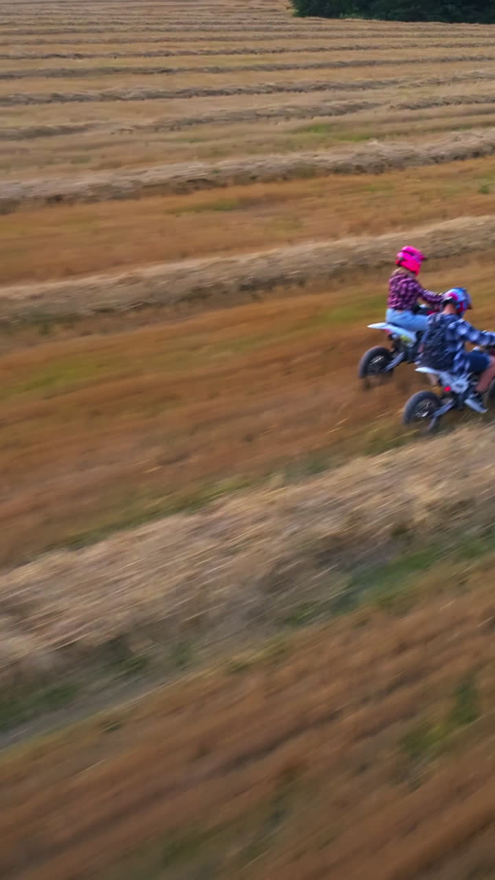 Young couple riding motorcycles close to each other. People having fun in the field with mowed grass at setting sun. View from above. Vertical video