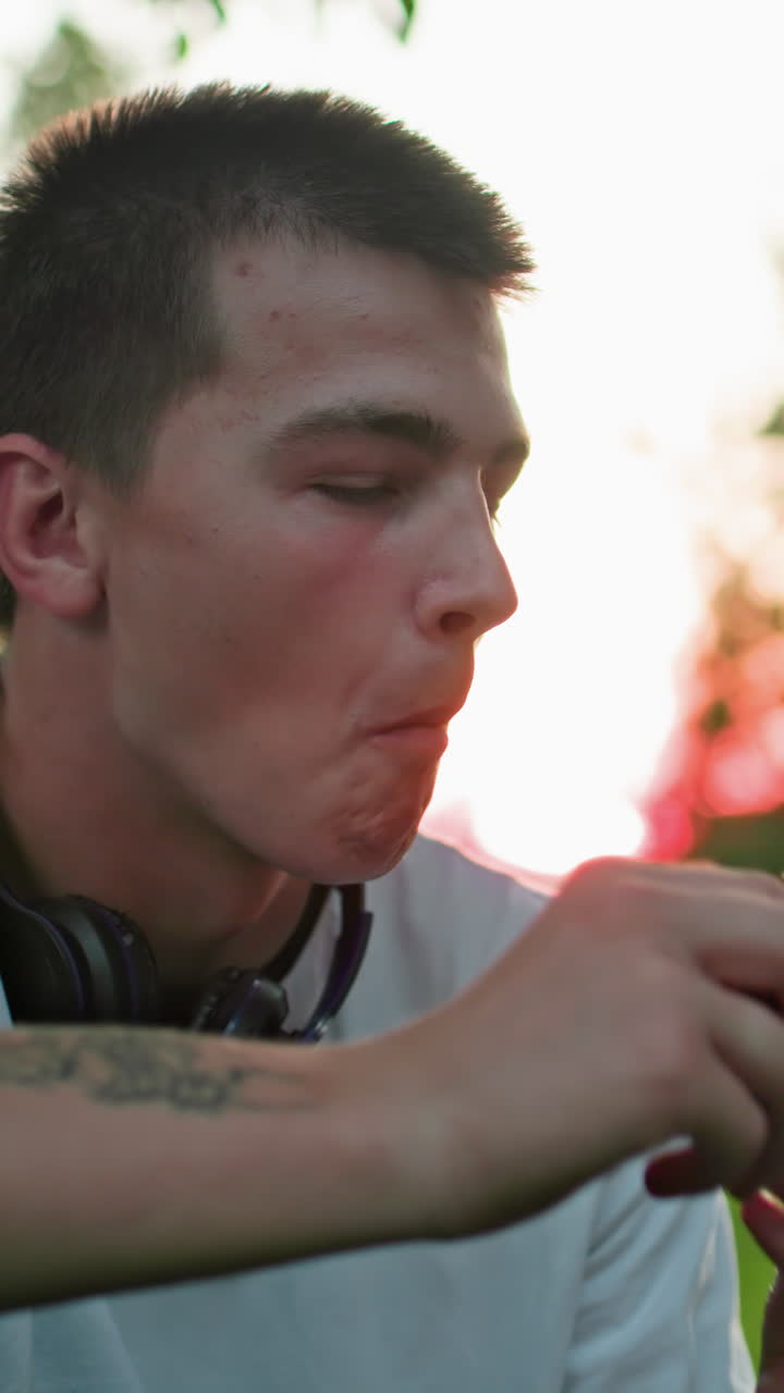 문신한 손을 가진 남자가 으로 토스트를 하는 동안 미소 짓고, 배경에서 을 들고 있는 다른 사람의 부분적인 시각으로, 빛은 나무를 통해 필터링됩니다.