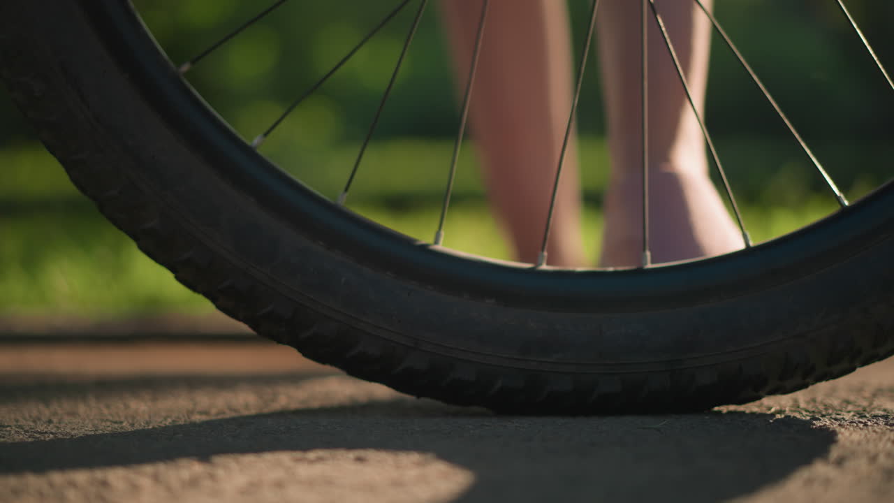 Close-up of individual legs in pink sneakers as they kick a bicycle tire to check air pressure, lush greenery and trees create a vibrant background, casting playful shadows on the ground