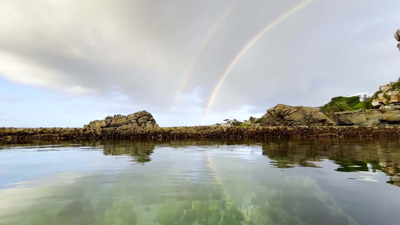 Klares Wasser in einem Gezeitenbecken mit einem doppelten Regenbogen am Himmel an einem bewölkten Morgen.