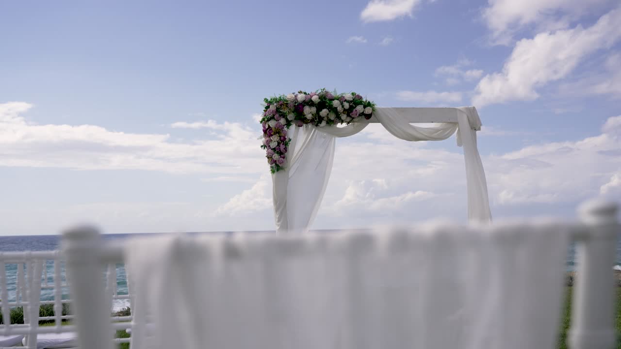 Allestimento per una cerimonia nuziale in riva all'oceano. Un arco floreale è pronto per la coppia. Sedie bianche allineate di fronte, dove gli ospiti si siederanno. Il cielo è limpido e luminoso.