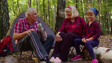 Senior-grandmother-grandfather-granddaughter-telling-funny-stories-fairy-tales-over-campfire-in-wood