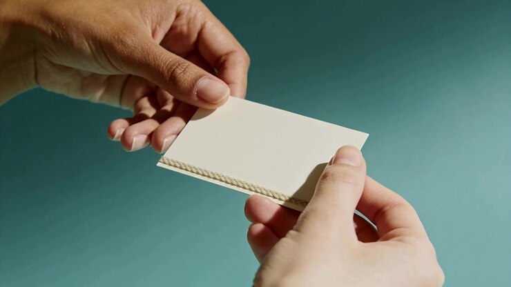 Two hands holding and tilting blank cream card with braided trim on teal background, AI-generated