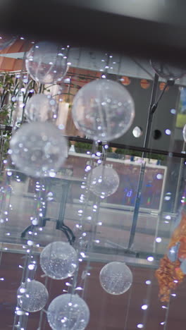 primer plano de decoraciones festivas con esferas translúcidas suspendidas con luces brillantes, reflejos borrosos de personas moviéndose a través de superficies de vidrio y vegetación vibrante