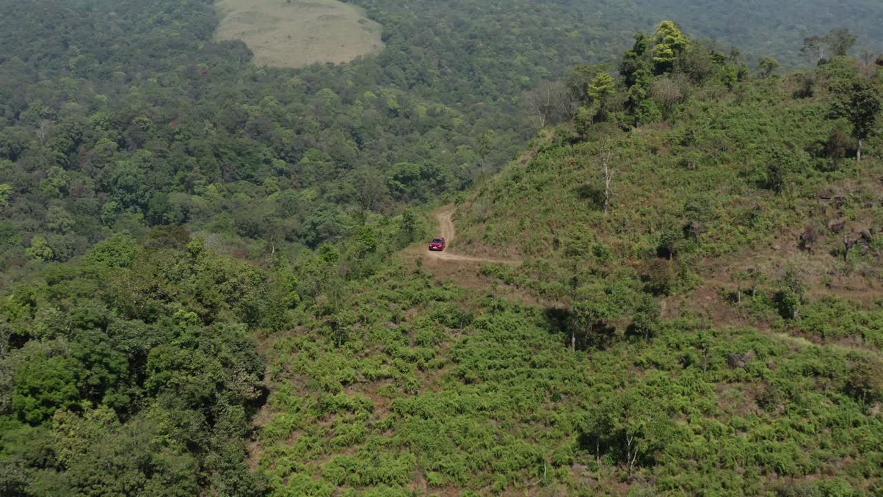 Drone trækker sig tilbage fra en rød bil, der kører op ad en snoet grusvej gennem tropiske bakker, hvilket afslører et tæt skovkronetag og ryddede bjergskråninger i Wayanad, Kerala, Indien