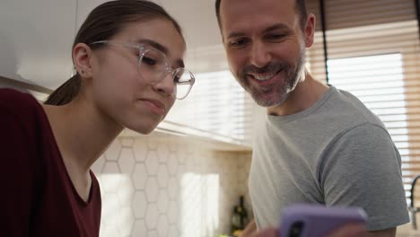 Caucasian-father-with-teenage-daughter-looking-at-mobile-phone-in-kitchen-while-cooking