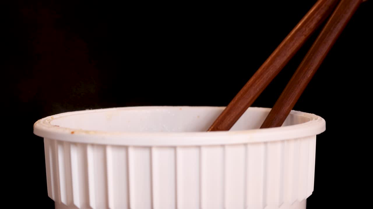 A hand skillfully lifts cooked instant noodles from a white cup using wooden chopsticks against a dark background, with soft, even lighting and close-up framing