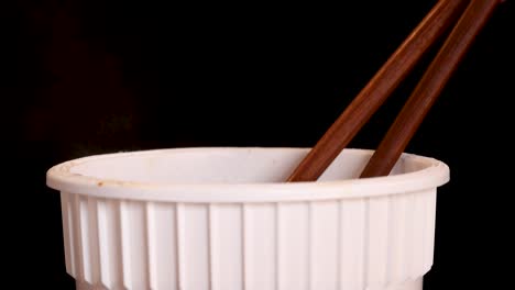 a hand skillfully lifts cooked instant noodles from a white cup using wooden chopsticks against a dark background, with soft, even lighting and close-up framing