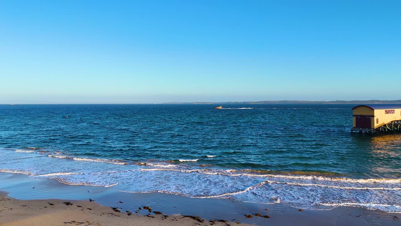 Calm ocean waves gently lap against the sandy shore near a pier under clear blue skies at Bellarine Peninsula