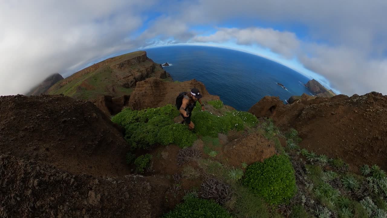 Solo hiker walking up Espigao Amorelo mountain in Madeira with a 360 action camera. Beautiful green cliffside with some clouds and blue sky. His friend is waiting at the top.