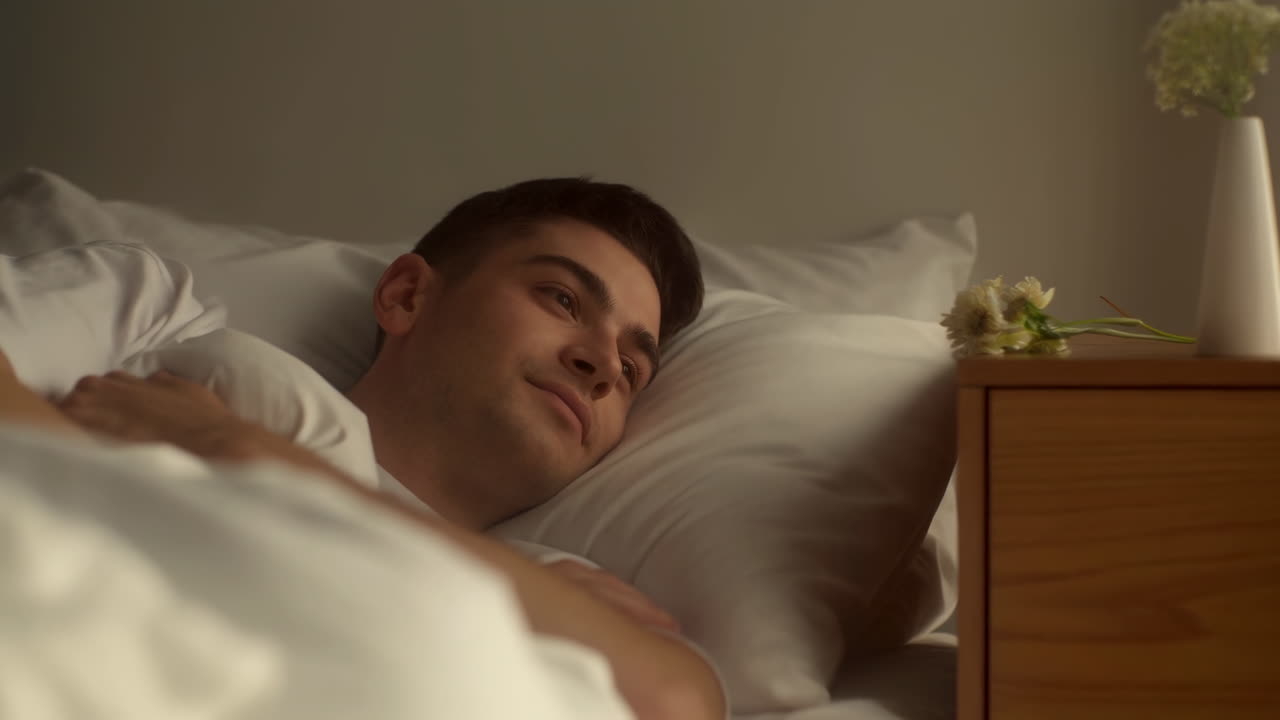Ung mand vågner i seng, blødt morgenlys, hvidt sengetøj, afslappet smil, natbord med vase