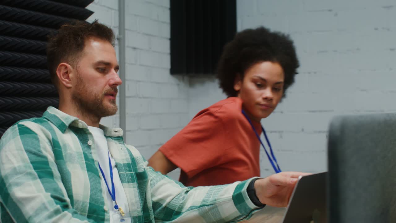 A man and woman work at laptops, sitting at one work desk in a office
