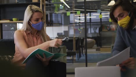 caucasian business people wearing face masks sitting going through paperwork in modern office