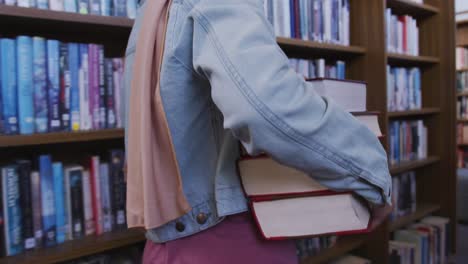 asian female student walking between bookshelves and carrying a stack of books