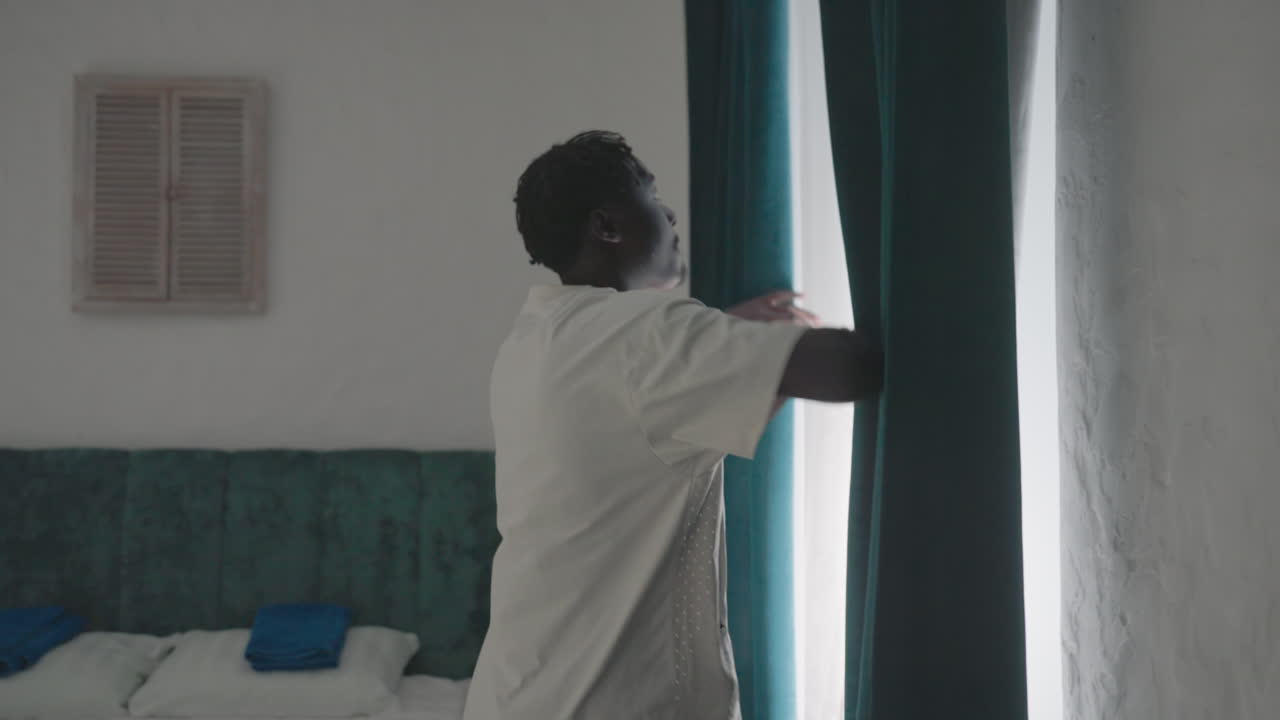 Man in white t-shirt standing in minimalist bedroom, gazing outside through dark green curtains, with green headboard, neatly made bed, and decorative window shutters in the background