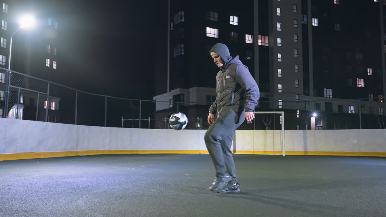 atleta hábilmente joggling fútbol en el campo al aire libre urbano por la noche, establecido contra un telón de fondo de edificios de gran altura iluminados y un cielo nocturno despejado