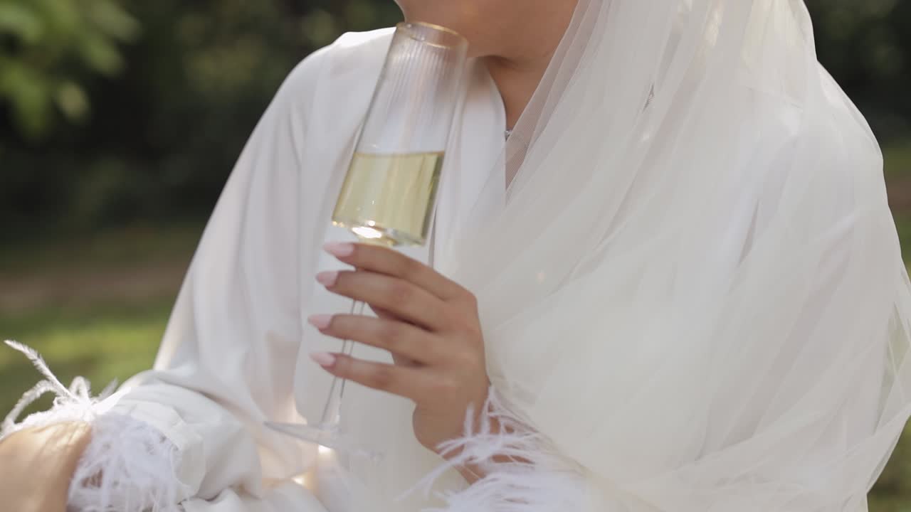 Sposa in abito boudoir seduta in un giardino mattutino, beve champagne, donna in camicia da notte e velo.