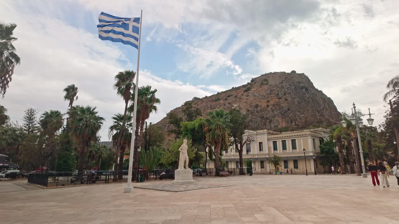 Greece national flag flies in city paved square under Palamidi fortress