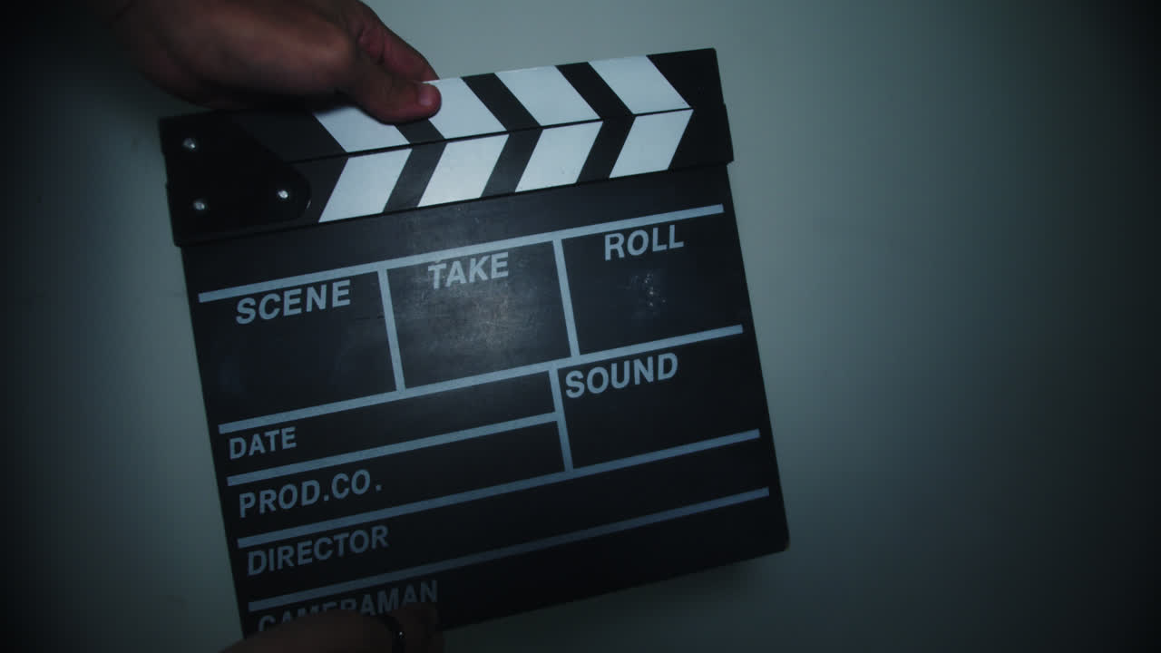 Beautiful slow motion shot of a man opening and closing a film clapperboard to announce a shot and give action to the shoot. It is in a room with a white wall and direct light.