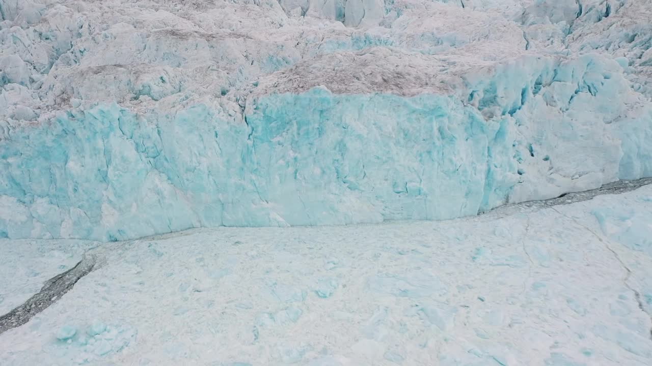 Aerial drone view of a massive glacier wall with vivid blue ice and rugged textures in Greenland, capturing the raw beauty and power of the Arctic’s frozen landscape