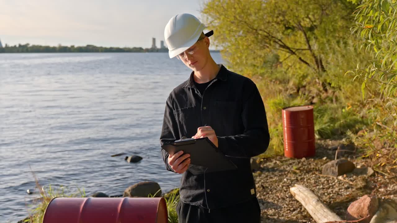 Young inspector writes on clipboard while surveying oil drum near polluted lakeshore