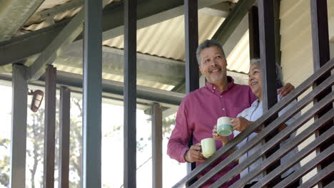 Feliz-Pareja-Birracial-De-Ancianos-Abrazándose-En-El-Balcón,-Hablando-Y-Bebiendo-Té