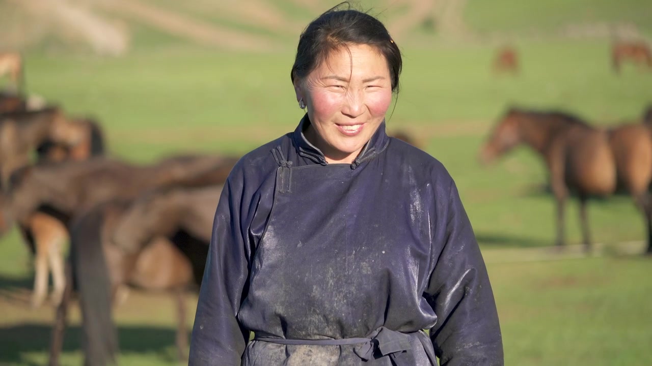 馬がいる牧草地に立つ民族衣装のモンゴル人女性