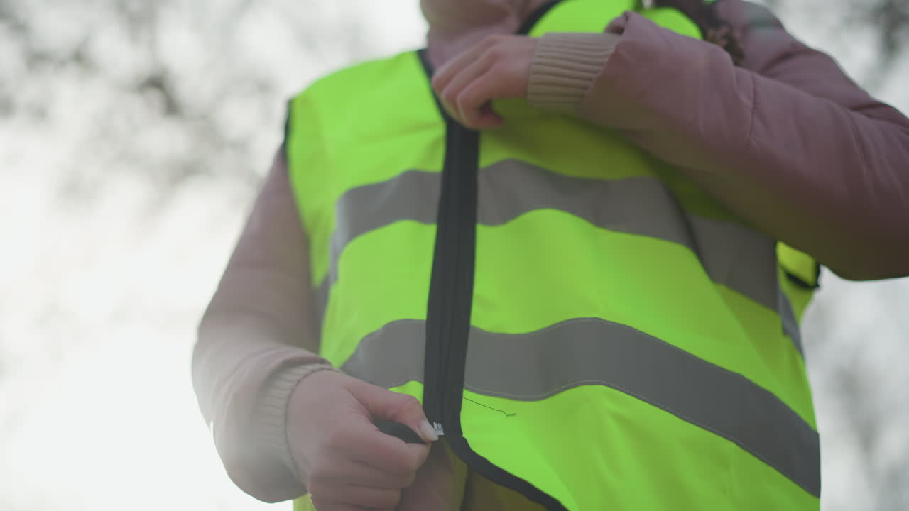 Close-up of woman in pink winter jacket fastening zipper on bright yellow reflective safety vest outdoors in daylight, preparing for work, volunteer activity, or safety duty requiring visibility