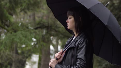 portrait of the worried, nervous young woman in a black jacket holding the black umbrella in the park