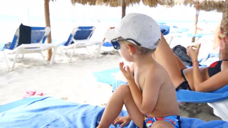 Niño-Sentado-En-La-Tumbona-En-La-Playa-Juega-Con-Su-Sombrero-Y-Gafas-De-Sol-Sacándolos-Y-Poniéndolos-Varias-Veces