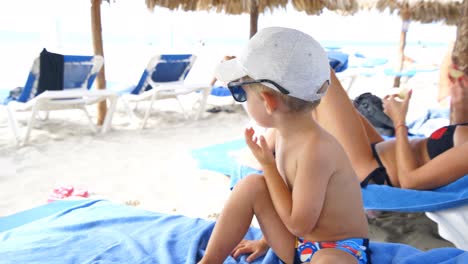 niño sentado en la tumbona en la playa juega con su sombrero y gafas de sol sacándolos y poniéndolos varias veces