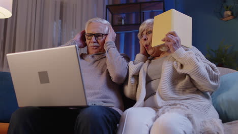 Senior-grandparents-couple-reading-book,-using-laptop-pc-on-couch-in-night-living-room-at-home