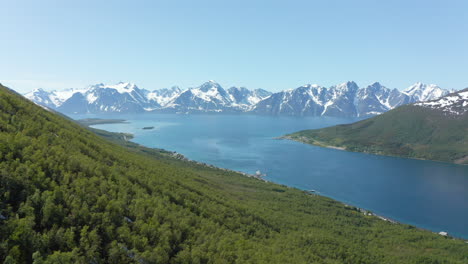 Aerial-view-over-forest,-towards-the-arctic-ocean,-snowy-Lyngen-alpine-mountains-in-the-background,-sunny,-summer-day,-in-Rotsund,-Troms,-Nordland,-Norway---Dolly,-drone-shot