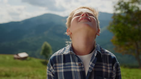 little boy enjoying sunlight raising head to sky in summer mountains close up.