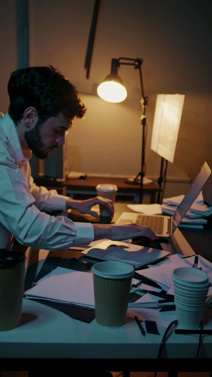 A dimly lit video scene captures a man working late at a cluttered desk, viewed from a side angle