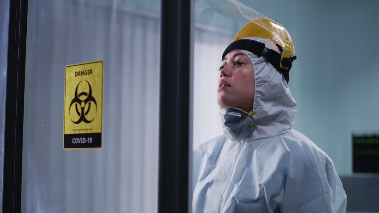 A healthcare worker in a hazmat suit and PPE, looking exhausted next to a COVID-19 biohazard sign