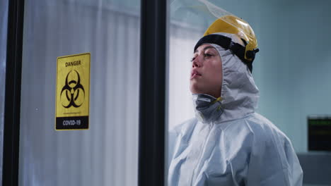 a healthcare worker in a hazmat suit and ppe, looking exhausted next to a covid-19 biohazard sign