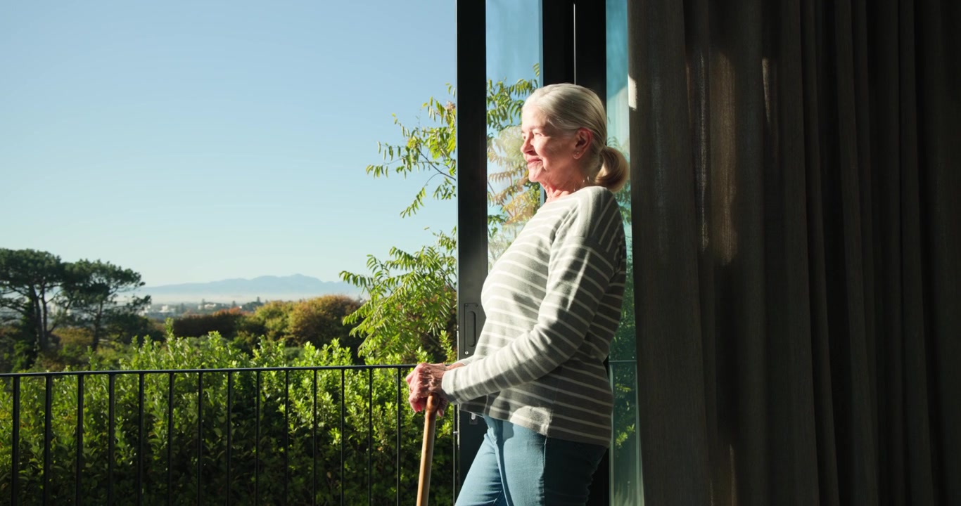 Donna anziana in piedi davanti a una porta scorrevole che tiene un bastone, con la luce del sole che le illumina il viso, mentre si gira, spazio per il testo.