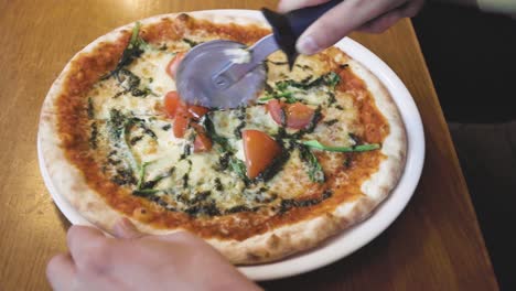 Close-up-of-pizza-being-cut-by-a-man-with-a-pizza-cutter-into-slices-seen-from-above-on-a-wooden-table