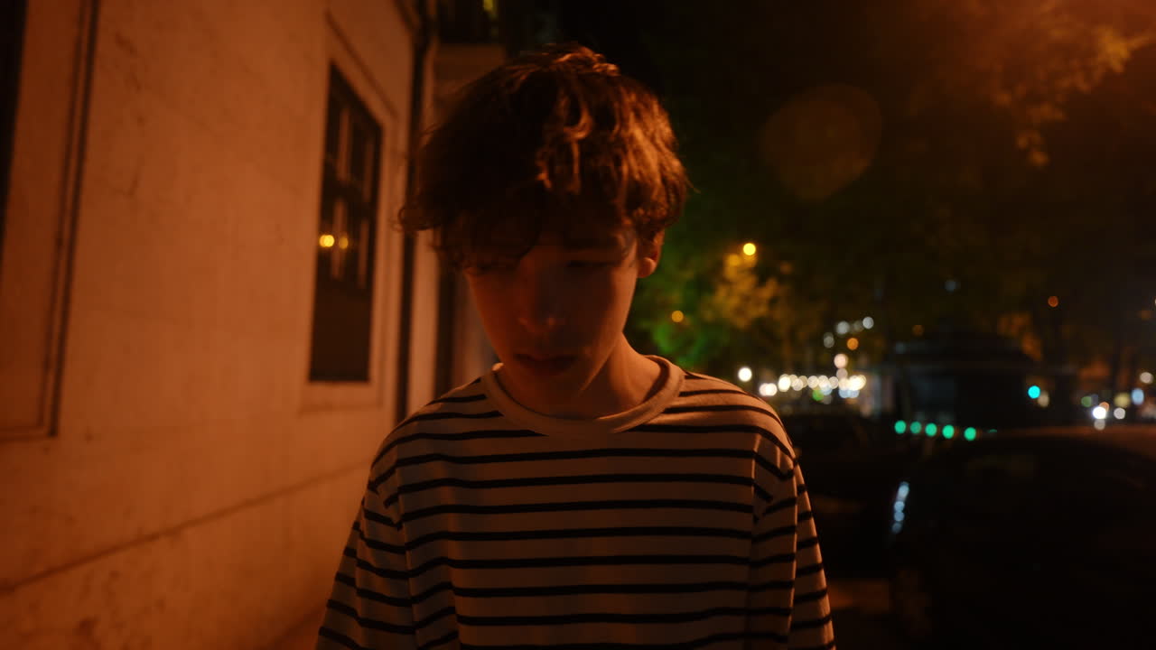 Teenage boy walking alone through dimly lit urban street at night, passing parked cars and shadowy buildings under soft street lamp illumination, embodying quiet contemplation